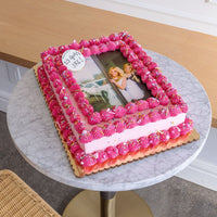A Classic Sheet Cake by Jenna Rae Cakes with pink frosting and piped borders displays a printed photo of two smiling women. A white plaque on top reads 50 years, NRC! The cake rests on a marble table.