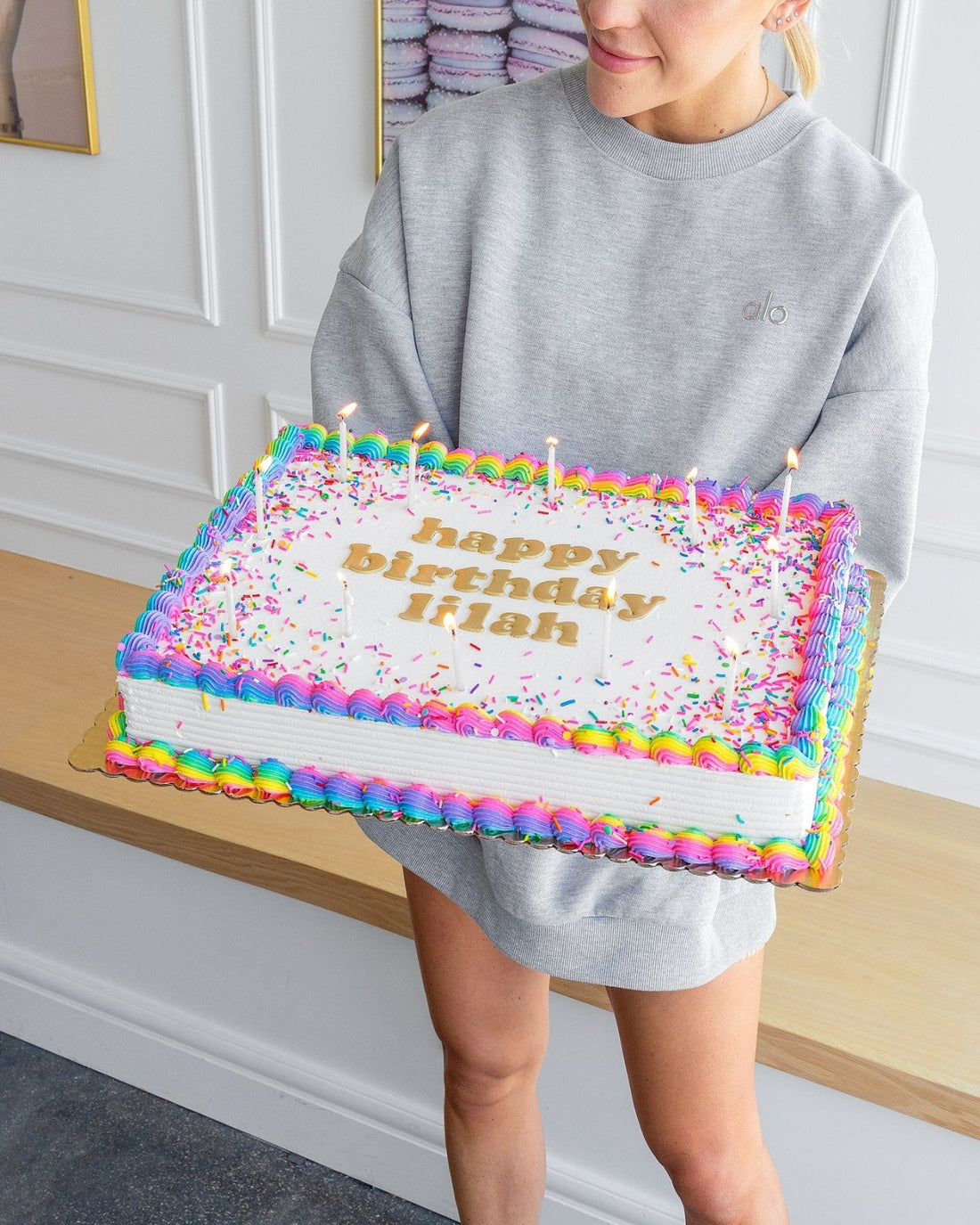 A person in a gray sweatshirt holds a Jenna Rae Cakes Classic Sheet Cake decorated with colorful icing, sprinkles, candles, and happy birthday Brian written on top.