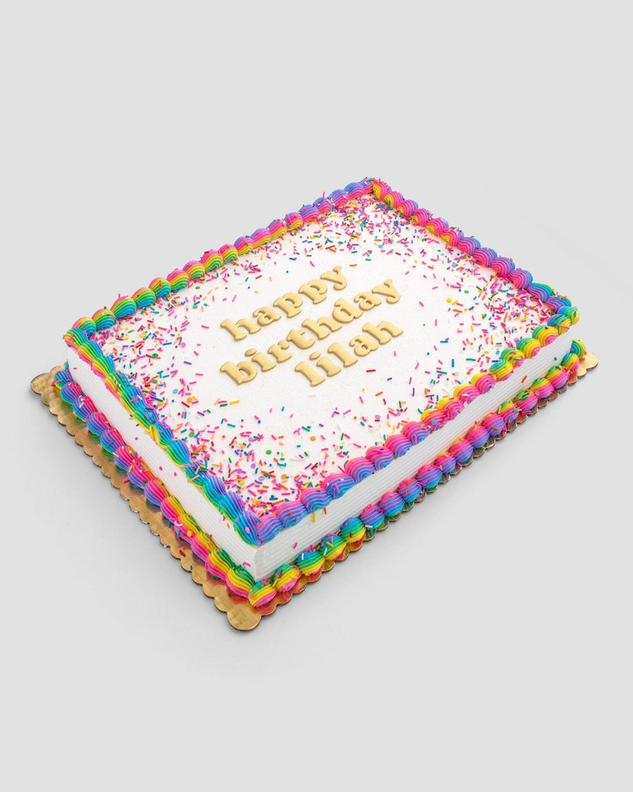 The Classic Sheet Cake by Jenna Rae Cakes features rainbow icing borders, rainbow sprinkles, and happy birthday Hilah in yellow on top. Presented on a gold board with a plain light background.
