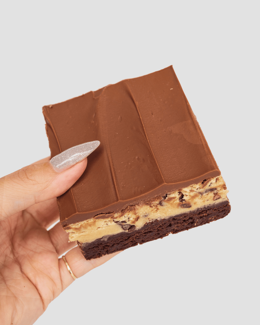 A hand with glittery nail polish holds a layered dessert bar featuring a brownie base, a cookie dough center with chocolate chips, and a smooth chocolate topping.