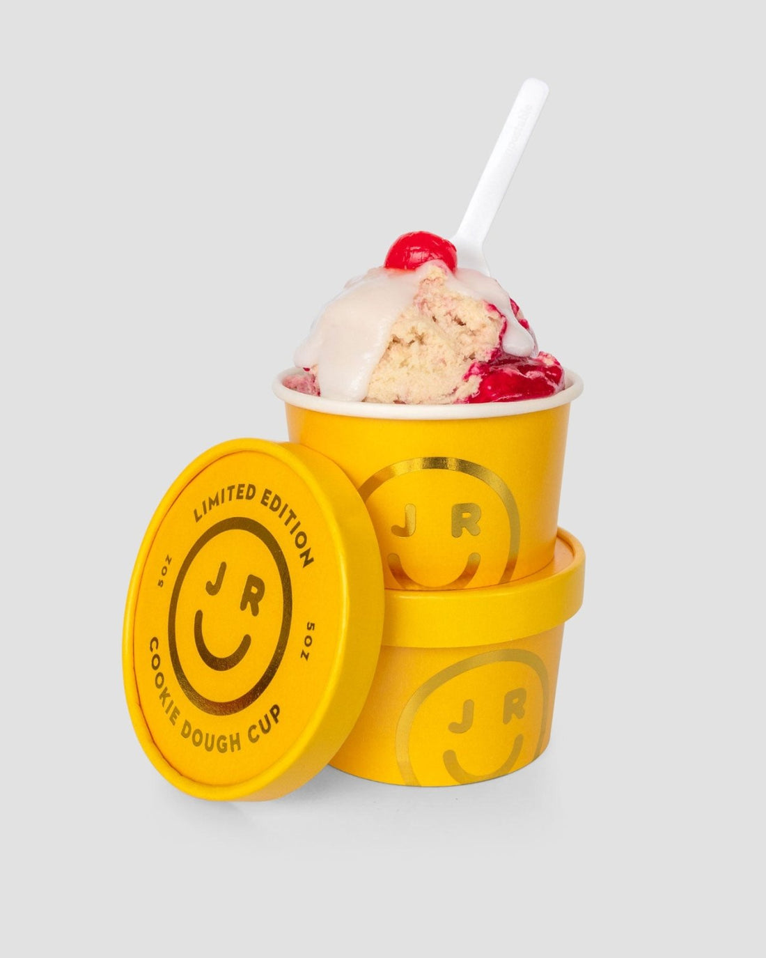 A yellow Jenna Rae Cakes Cookie Dough Cup labeled Limited Edition shows a smiley face and is filled with cookie dough, red syrup, white icing, a cherry, and a white spoon. A second identical cup and its lid rest nearby on a gray background.