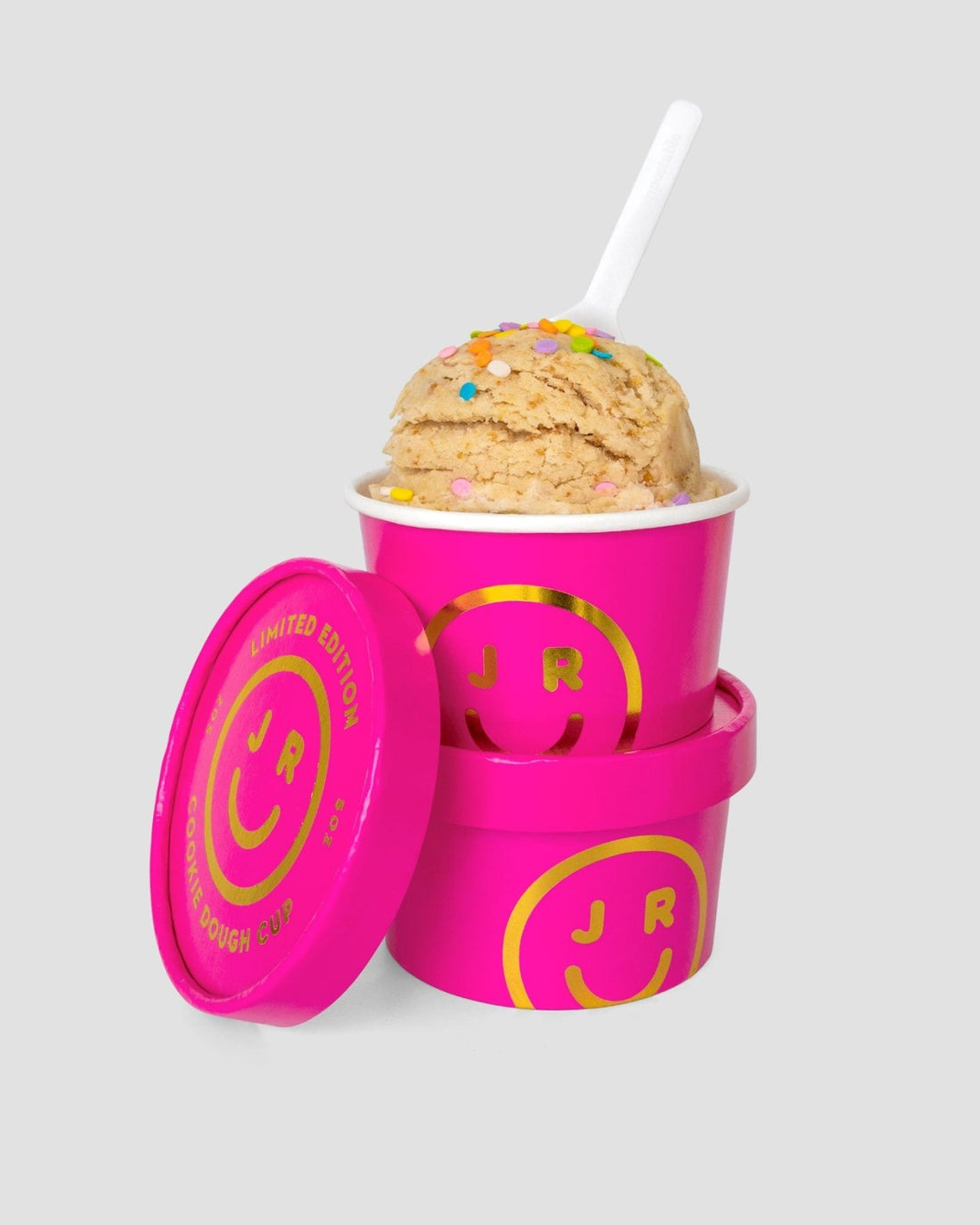 Two Cookie Dough Cups from Jenna Rae Cakes, each in a bright pink container with a yellow smiley face and JR logo—one topped with colorful sprinkles and a white spoon, the other stacked below with its lid off.