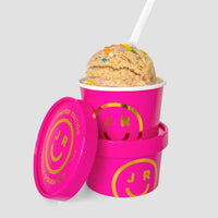Two Cookie Dough Cups from Jenna Rae Cakes, each in a bright pink container with a yellow smiley face and JR logo—one topped with colorful sprinkles and a white spoon, the other stacked below with its lid off.