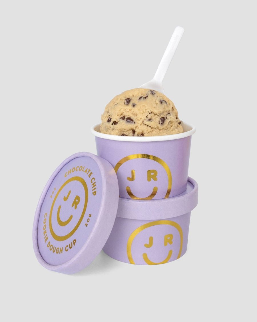 A scoop of Cookie Dough Cups by Jenna Rae Cakes in a purple cup with gold smiley face and JR branding, stacked on another matching cup, with a white spoon and matching lid nearby.