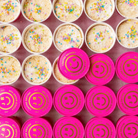 Rows of colorful Jenna Rae Cakes Cookie Dough Cups are arranged neatly. The top row shows open cups with confetti sprinkles, while the bottom rows have bright pink Limited Edition lids featuring a smiley face.