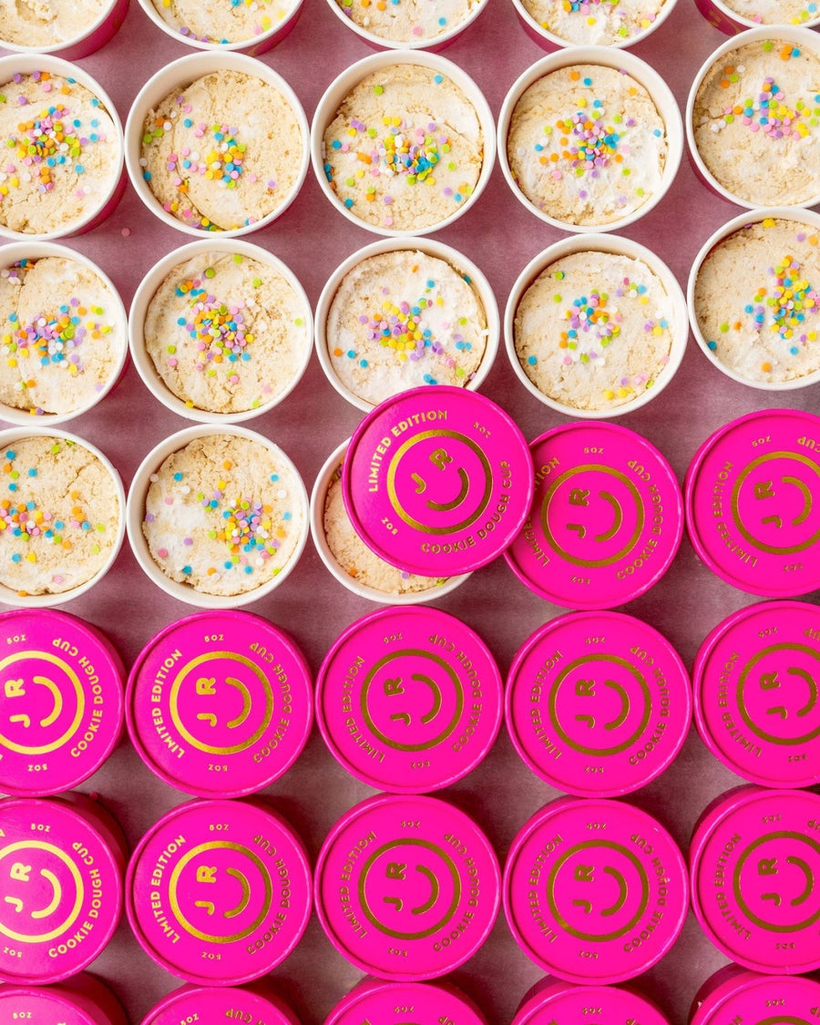 Rows of colorful Jenna Rae Cakes Cookie Dough Cups are arranged neatly. The top row shows open cups with confetti sprinkles, while the bottom rows have bright pink Limited Edition lids featuring a smiley face.