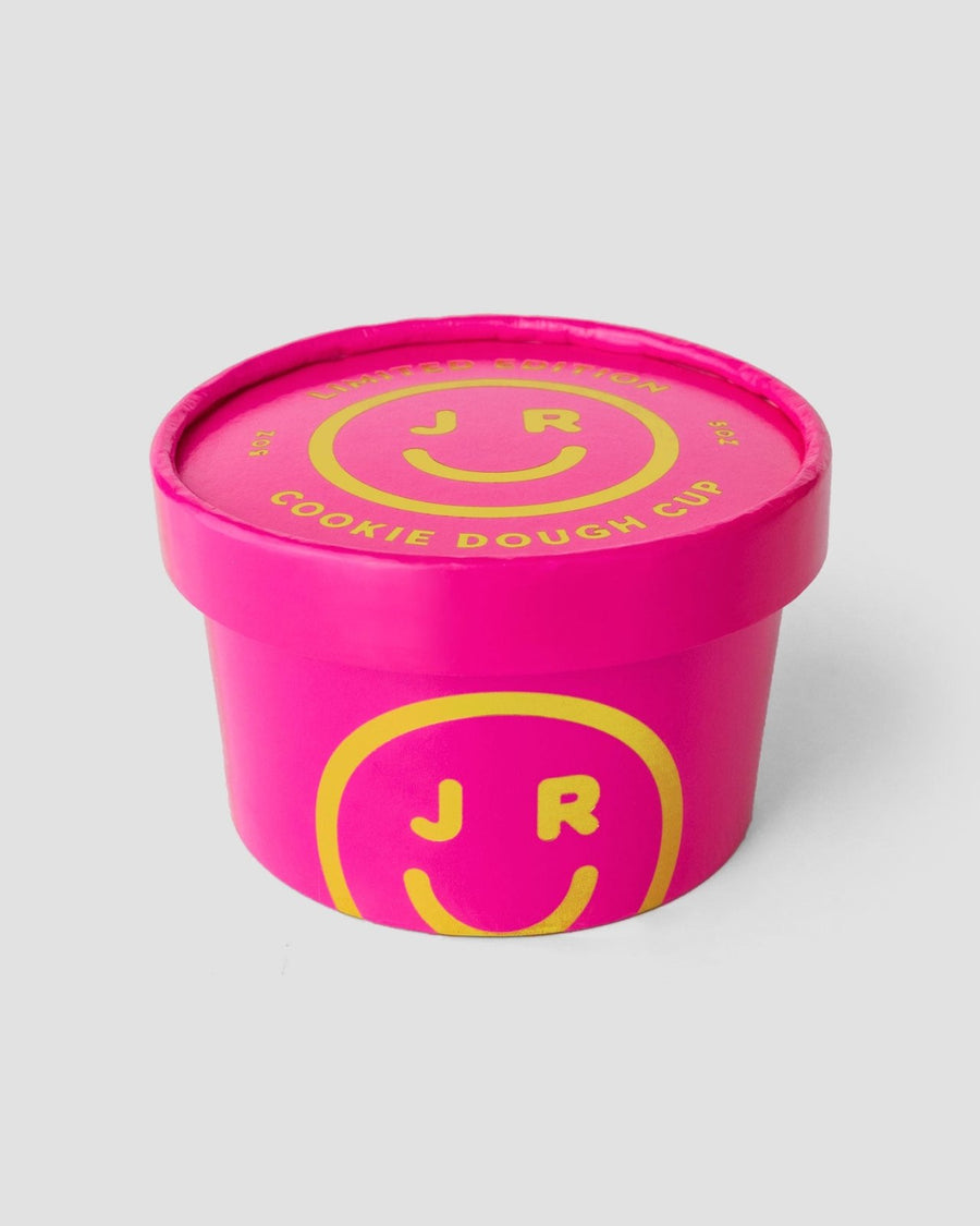 A bright pink Jenna Rae Cakes Cookie Dough Cup with a matching lid displays yellow smiley faces and JR lettering. The lid label says COOKIE DOUGH CUP in yellow. Packaging is shown on a plain, light gray background.