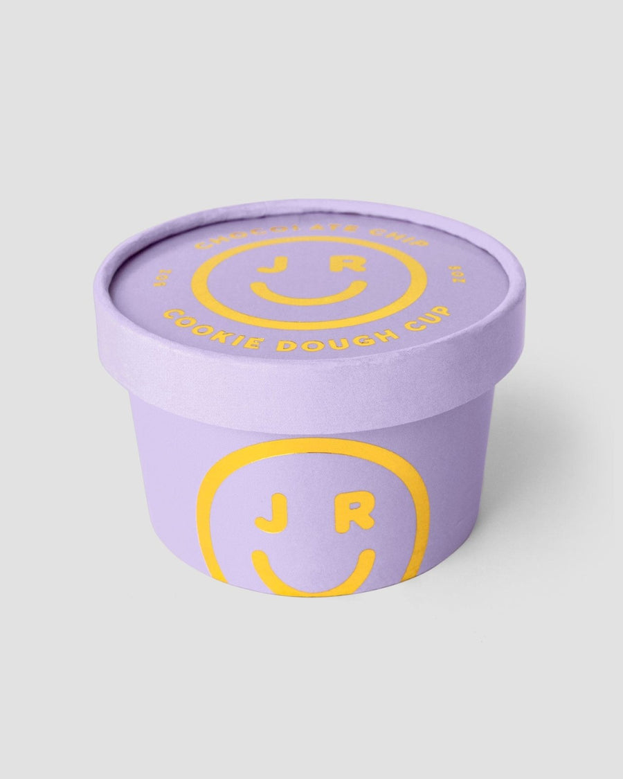 A lavender Jenna Rae Cakes Cookie Dough Cup with a matching lid, both featuring COOKIE DOUGH CUP in yellow and a smiley face with J R inside on the side and top.