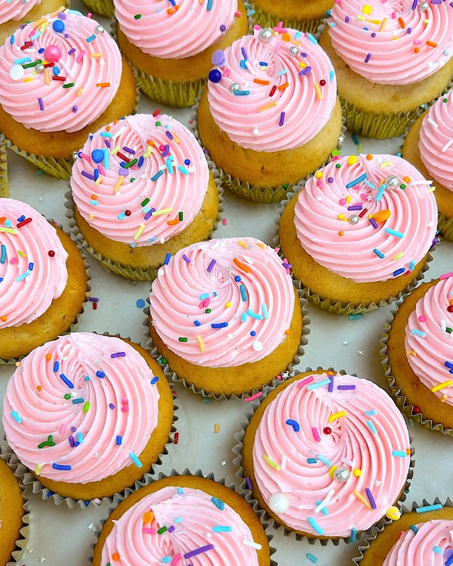 Rows of vanilla cupcakes with pink swirled frosting and colorful sprinkles, arranged on a white surface, by Jenna Rae Cakes for their Dessert Tables.