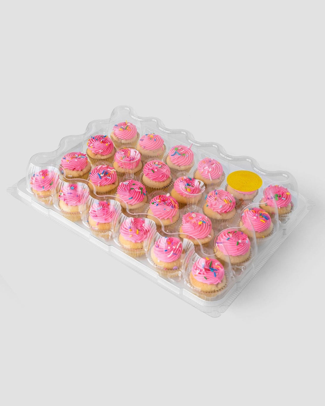 A clear plastic tray of 24 mini cupcakes with pink frosting and colorful sprinkles from Jenna Rae Cakes Dessert Tables line, featuring one empty slot covered by a yellow sticker, set against a plain light gray background.