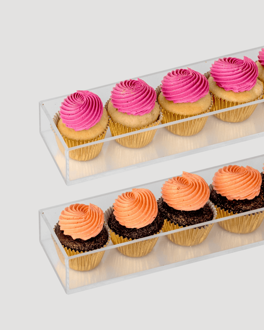 The Dessert Tables by Jenna Rae Cakes features two clear rectangular trays on a light gray background—one with four pink-frosted cupcakes, the other with four orange-frosted cupcakes topped with chocolate sprinkles.