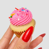 A hand with long, glossy red nails holds a vanilla cupcake with pink frosting and sprinkles from the Dessert Tables collection by Jenna Rae Cakes, set against a plain light background.
