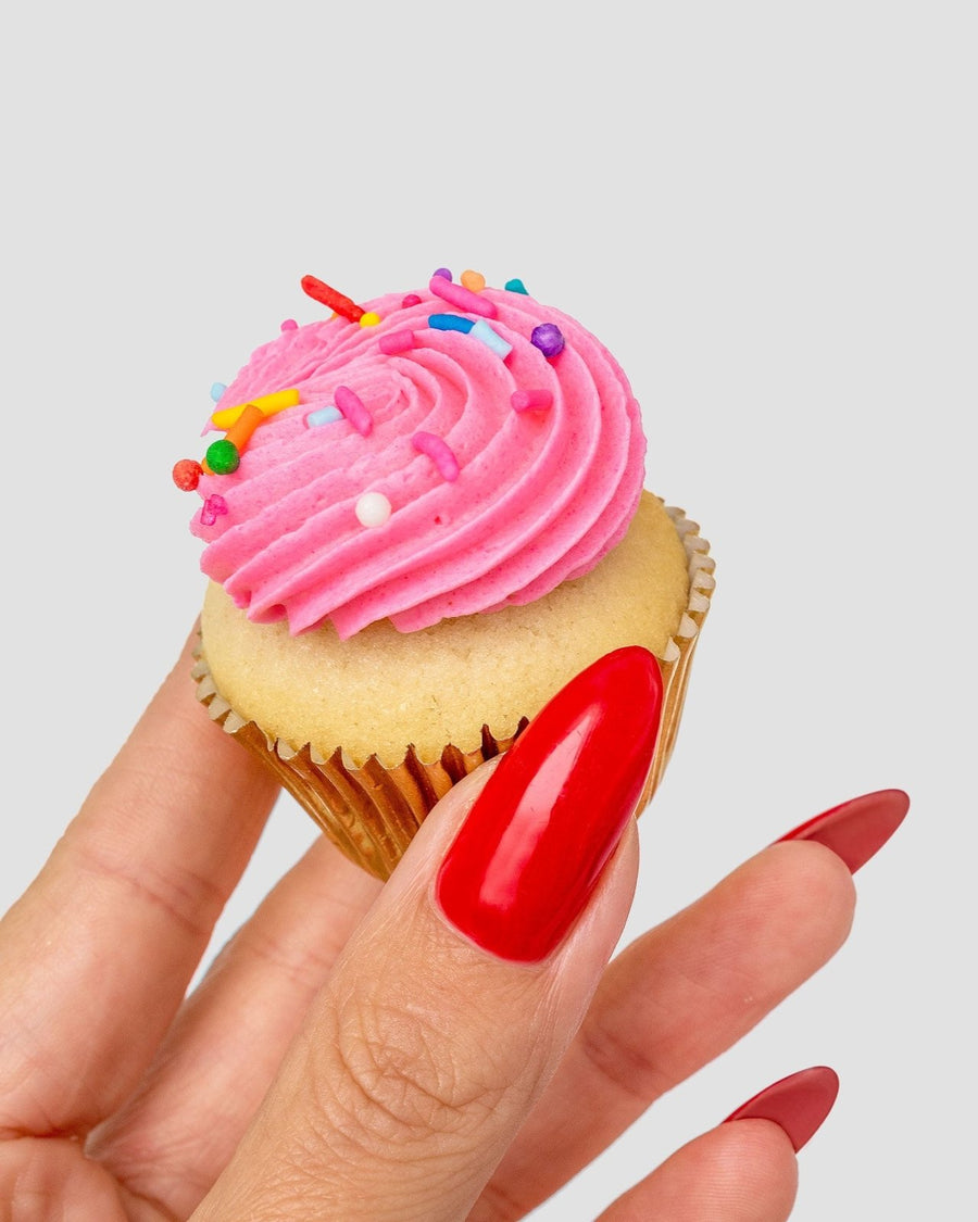 A hand with long, glossy red nails holds a vanilla cupcake with pink frosting and sprinkles from the Dessert Tables collection by Jenna Rae Cakes, set against a plain light background.