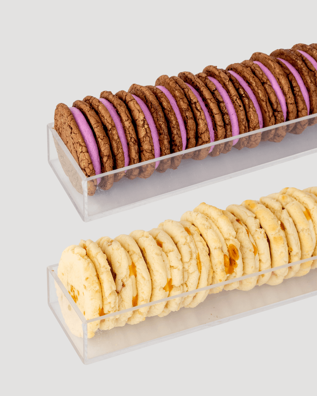 Two rows of Dessert Tables sandwich cookies by Jenna Rae Cakes in clear trays: the top row has chocolate cookies with purple filling, while the bottom row features light cookies with orange bits. Both trays sit on a light gray background.