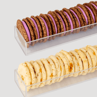 Two rows of Dessert Tables sandwich cookies by Jenna Rae Cakes in clear trays: the top row has chocolate cookies with purple filling, while the bottom row features light cookies with orange bits. Both trays sit on a light gray background.