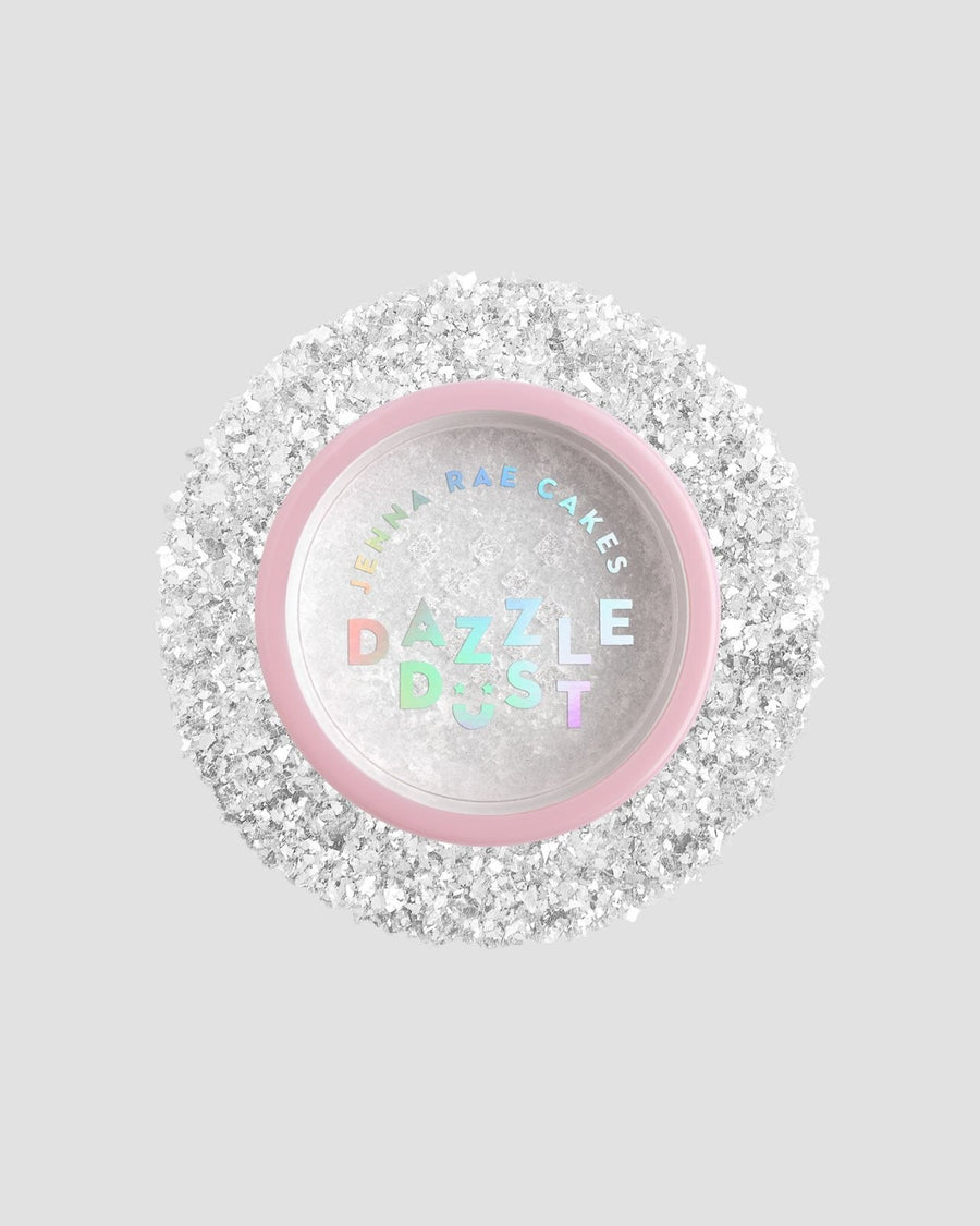 A small round container with a pink rim and clear lid labeled Jenna Rae Cakes Dazzle Dust Best Seller Trio, filled with fine, sparkly white glitter, sits on a light gray background.