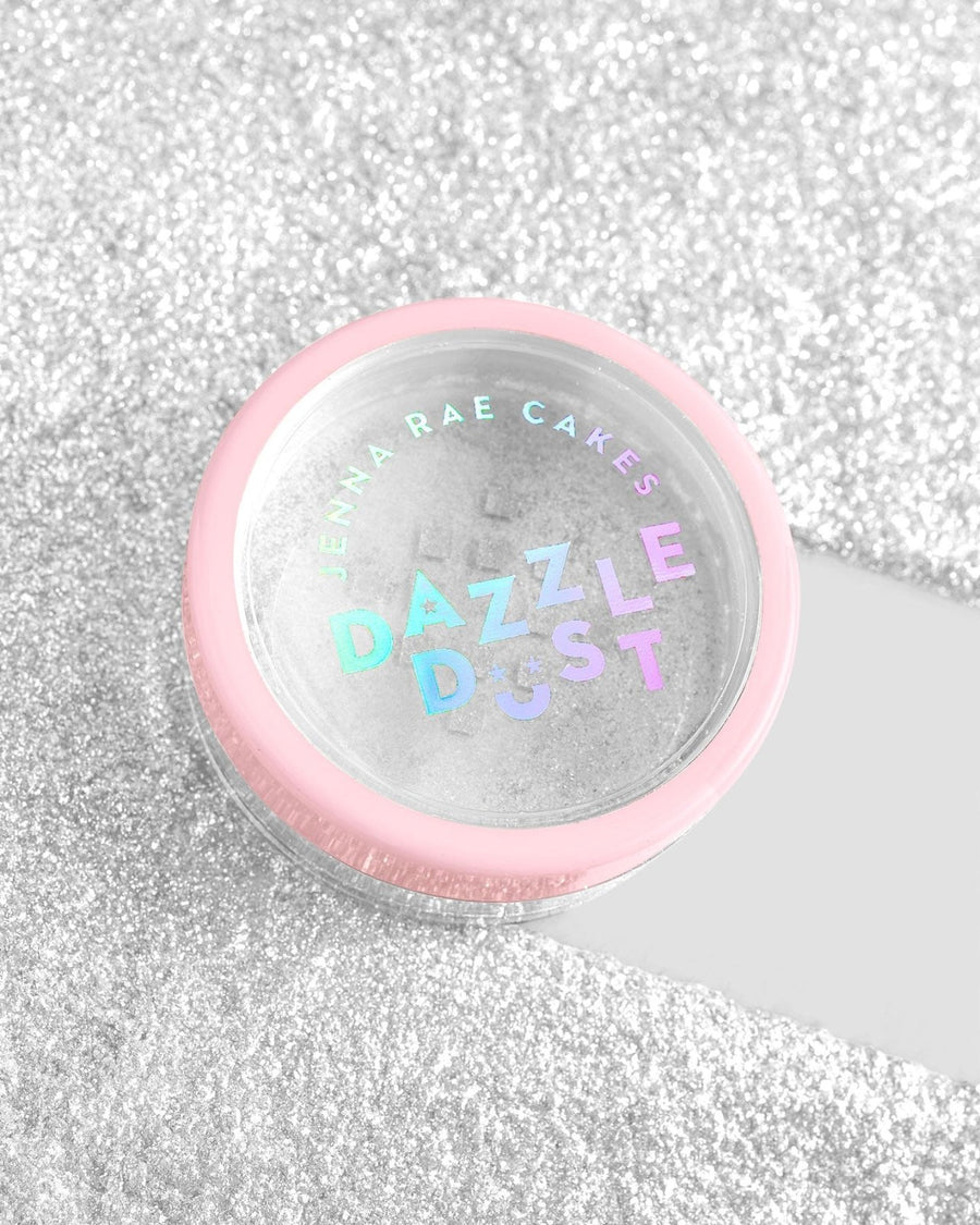 A small round container with a pink rim labeled Jenna Rae Cakes Dazzle Dust Best Seller Trio sits on a glittery silver surface, with its transparent lid revealing the shimmery powder inside.