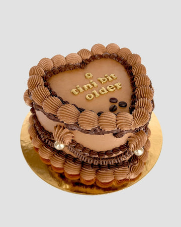 The Espresso Martini Heart Cake by Jenna Rae Cakes is a heart-shaped chocolate cake with decorative frosting, gold pearls, and “a tini bit older” written in gold letters on top, set on a gold cake board.