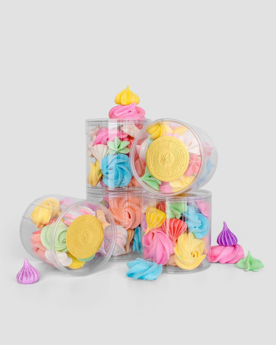 Jenna Rae Cakes Favours: Clear plastic jars filled with colorful meringue cookies in yellow, pink, blue, purple, and green are stacked on a light gray background, with some meringues scattered around.