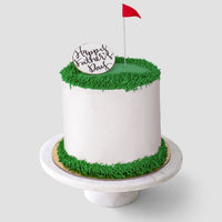 The Golf Ball Cake by Jenna Rae Cakes features white frosting, green grass-like icing on the edges, a small red flag topper, and a round “Happy Fathers Day” topper, all displayed on a white cake stand.
