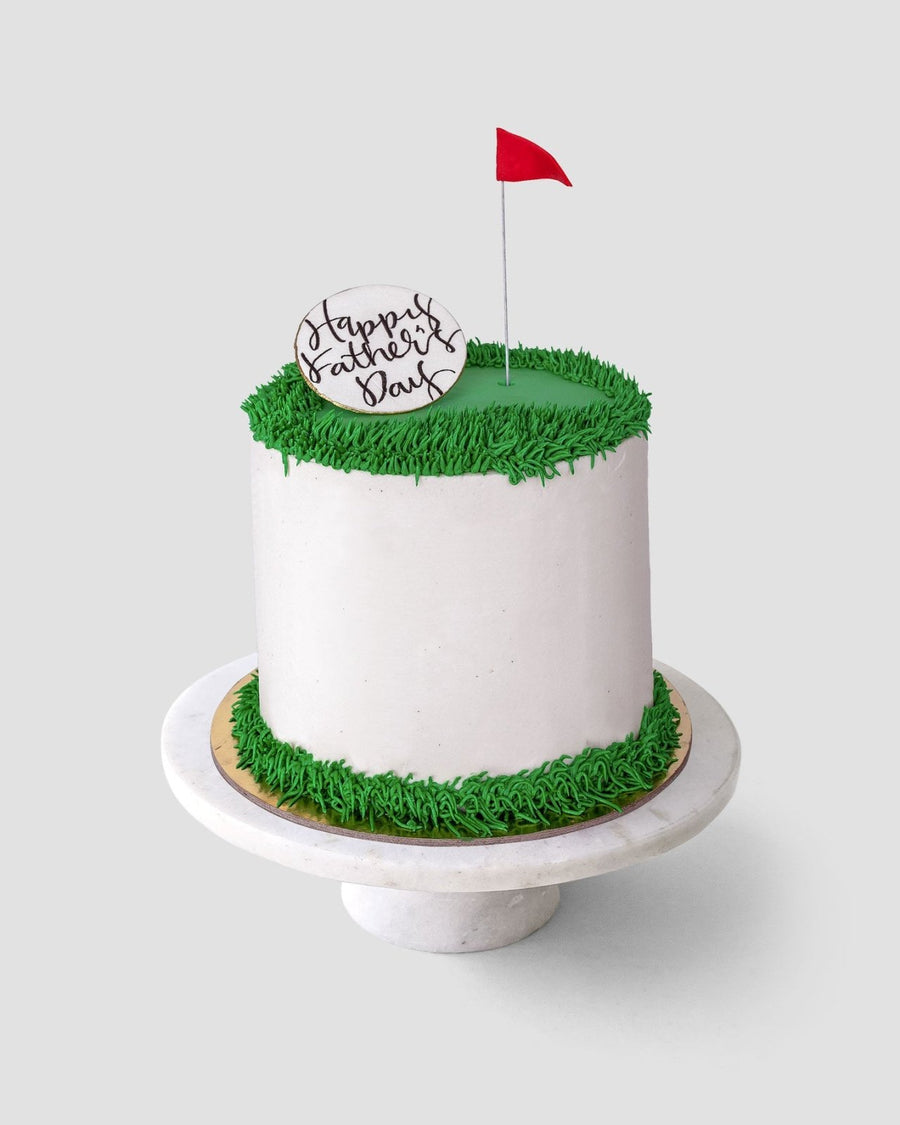 The Golf Ball Cake by Jenna Rae Cakes is a white frosted cake with green icing grass, topped with a red flag and a Happy Fathers Day plaque, displayed on a white cake stand.
