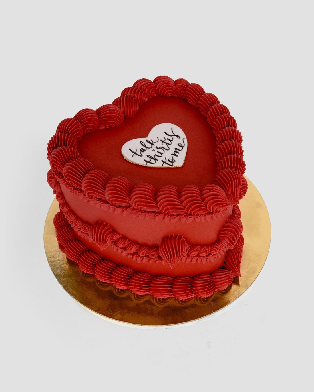 The Heart Cake by Jenna Rae Cakes is a heart-shaped red cake with decorative red frosting and a white heart on top reading talk strictly to me, presented on a round gold cake board.