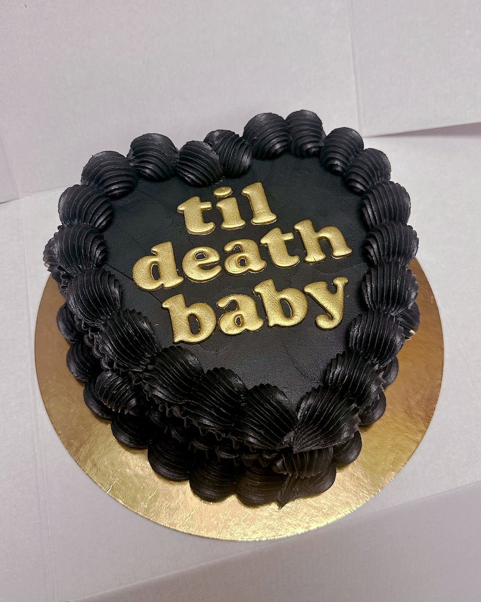 A heart-shaped black cake with thick, ruffled black frosting along the edges, featuring gold letters that spell out til death baby. The cake sits on a round golden board.