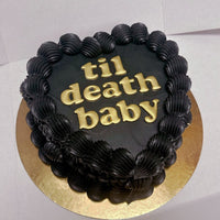 A heart-shaped black cake with thick, ruffled black frosting along the edges, featuring gold letters that spell out til death baby. The cake sits on a round golden board.
