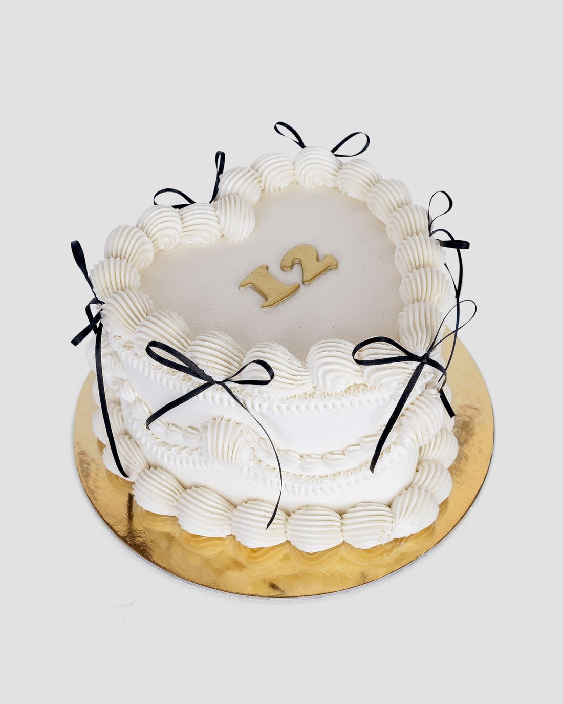 The Heart Cake by Jenna Rae Cakes is a round white frosted cake with black ribbon bows, piped icing, and a gold “12” on top. It sits on a gold cake board against a plain background.