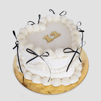 The Heart Cake by Jenna Rae Cakes is a round white frosted cake with black ribbon bows, piped icing, and a gold “12” on top. It sits on a gold cake board against a plain background.