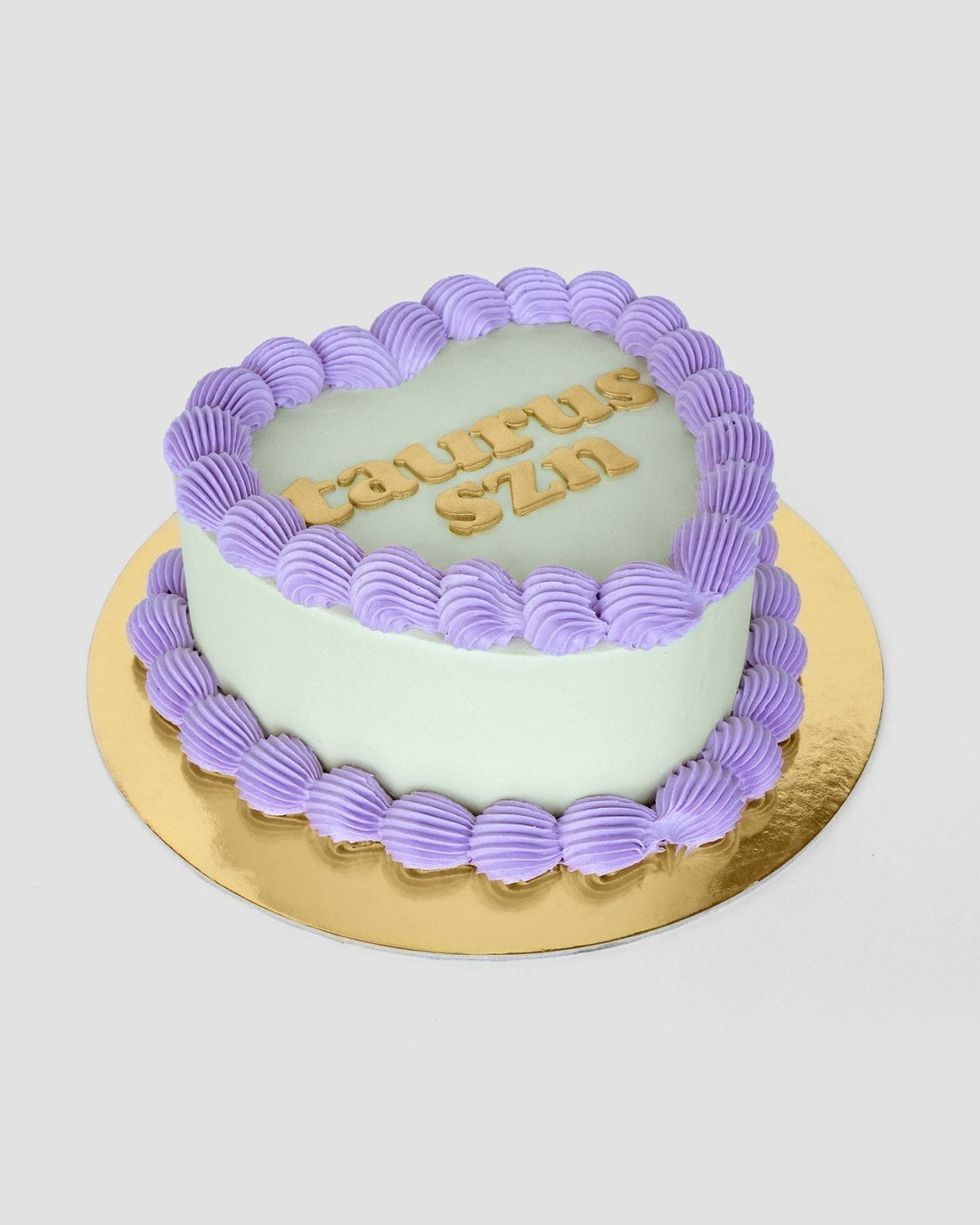 The Heart Cake by Jenna Rae Cakes features a heart shape with pale green icing, a purple frosting border, TAURUS SZN in yellow letters on top, all presented on a gold cake board.