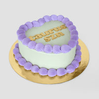 The Heart Cake by Jenna Rae Cakes features a heart shape with pale green icing, a purple frosting border, TAURUS SZN in yellow letters on top, all presented on a gold cake board.