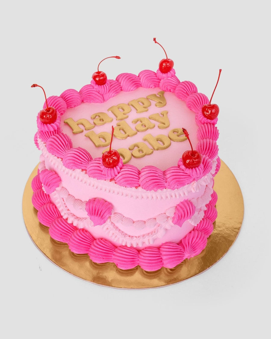 The Heart Cake by Jenna Rae Cakes is a pink, heart-shaped cake with piped pink icing, six cherries on top, and gold happy bday babe letters. It sits on a gold board against a plain light background.