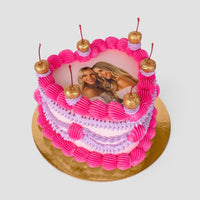 The Heart Cake by Jenna Rae Cakes features pink and purple frosting with five gold cherries on top. An edible photo of two smiling women is at the center. The cake sits on a gold board against a plain light background.