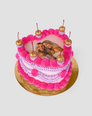 The Heart Cake by Jenna Rae Cakes features pink and purple frosting with five gold cherries on top. An edible photo of two smiling women is at the center. The cake sits on a gold board against a plain light background.