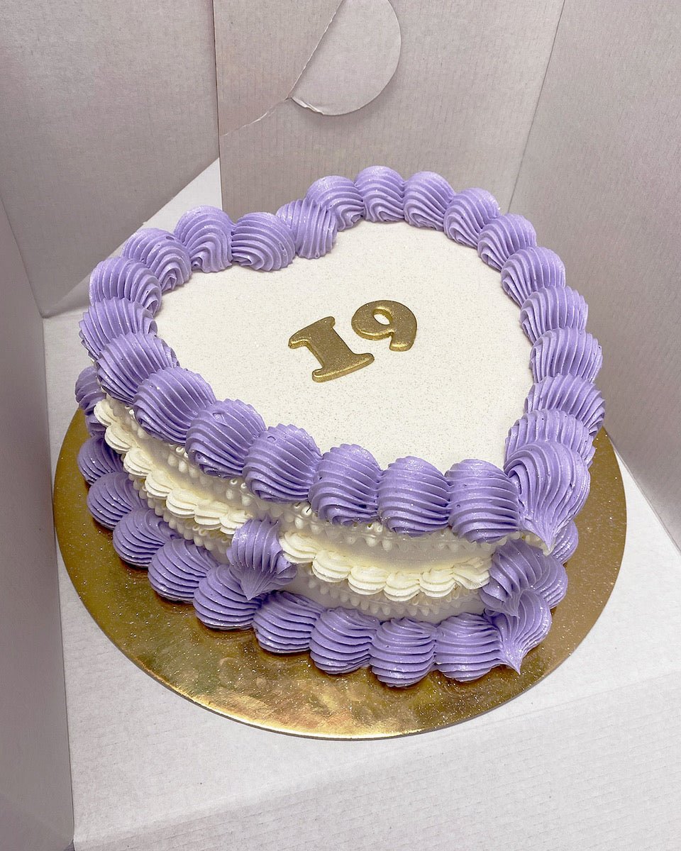 A heart-shaped cake with purple and white frosting, decorated with the number 19 in gold on top, placed on a gold cake board inside a white box.