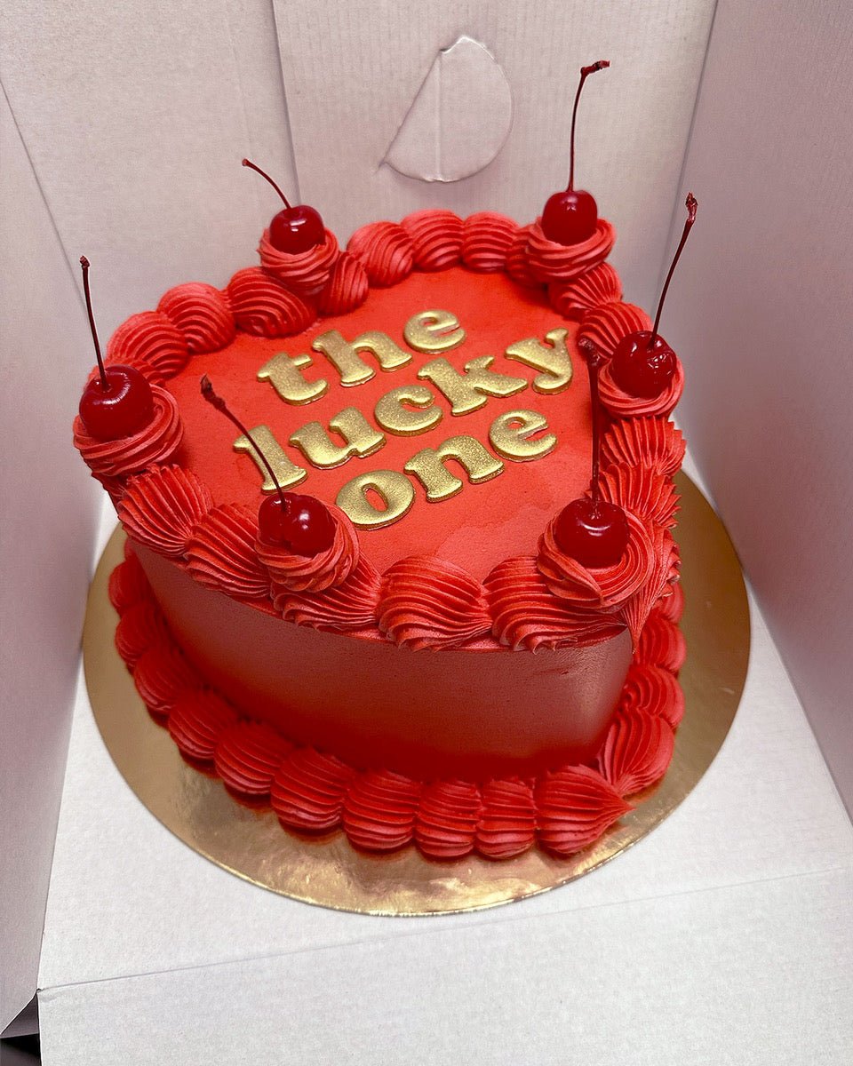 A heart-shaped red cake with red frosting, topped with five cherries and the words the lucky one written in gold letters, placed on a gold cake board inside a white box.