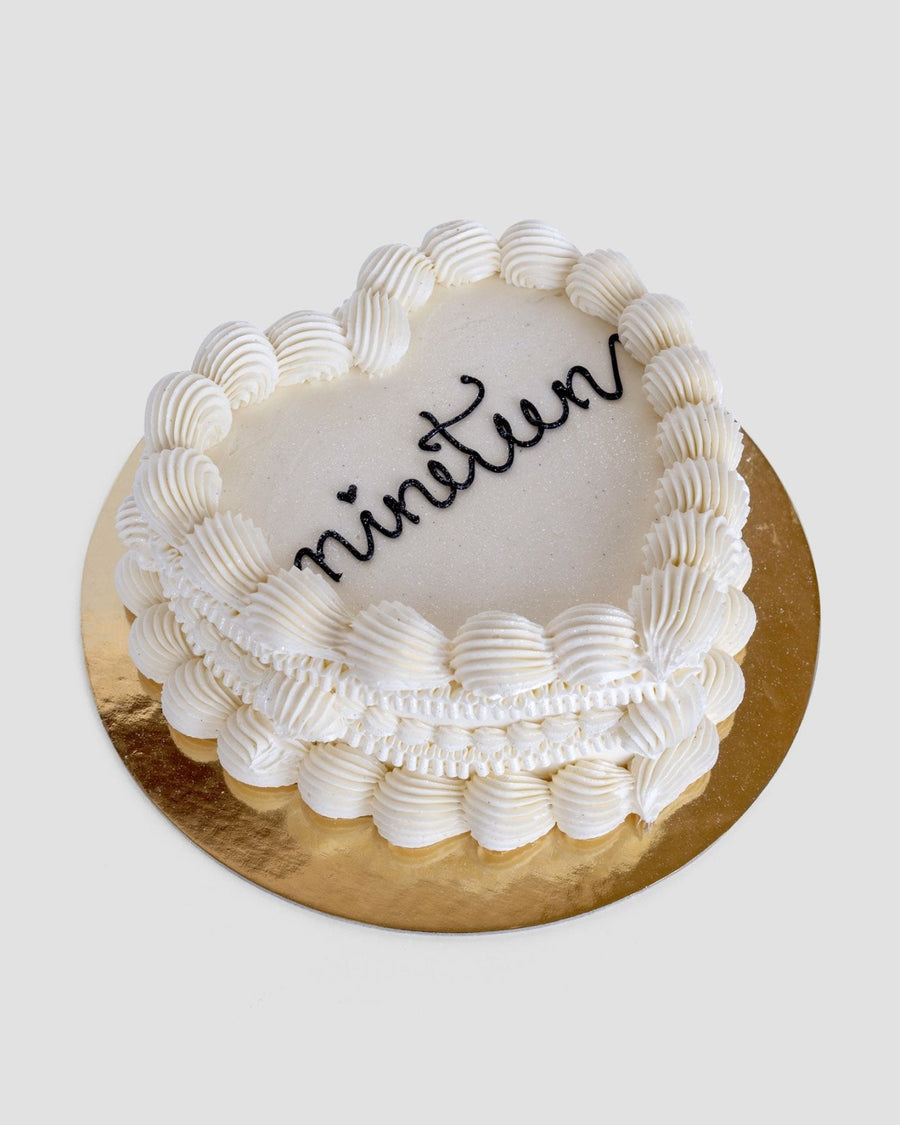 The Heart Cake by Jenna Rae Cakes is a white, heart-shaped cake with decorative icing borders and nineteen written in black icing, set on a round gold cake board.