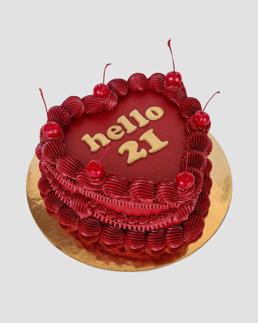 The Heart Cake by Jenna Rae Cakes is a heart-shaped red cake with decorative red frosting, six cherries on top, and “hello 21” in gold icing, presented on a round golden board.