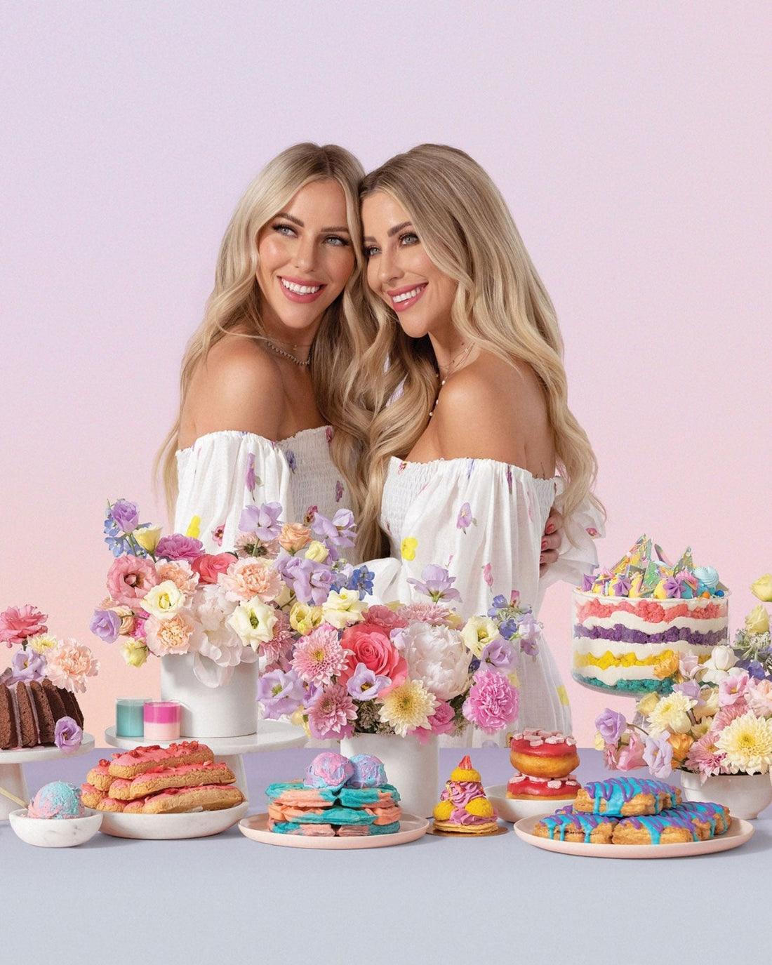 Two smiling women with long blonde hair in white off-shoulder dresses stand together among colorful flowers and vibrant pastries, showcasing the Jenna Rae Cakes at Home Cookbook by Jenna Rae Cakes against a soft pastel backdrop.