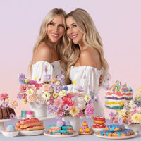 Two smiling women with long blonde hair in white off-shoulder dresses stand together among colorful flowers and vibrant pastries, showcasing the Jenna Rae Cakes at Home Cookbook by Jenna Rae Cakes against a soft pastel backdrop.