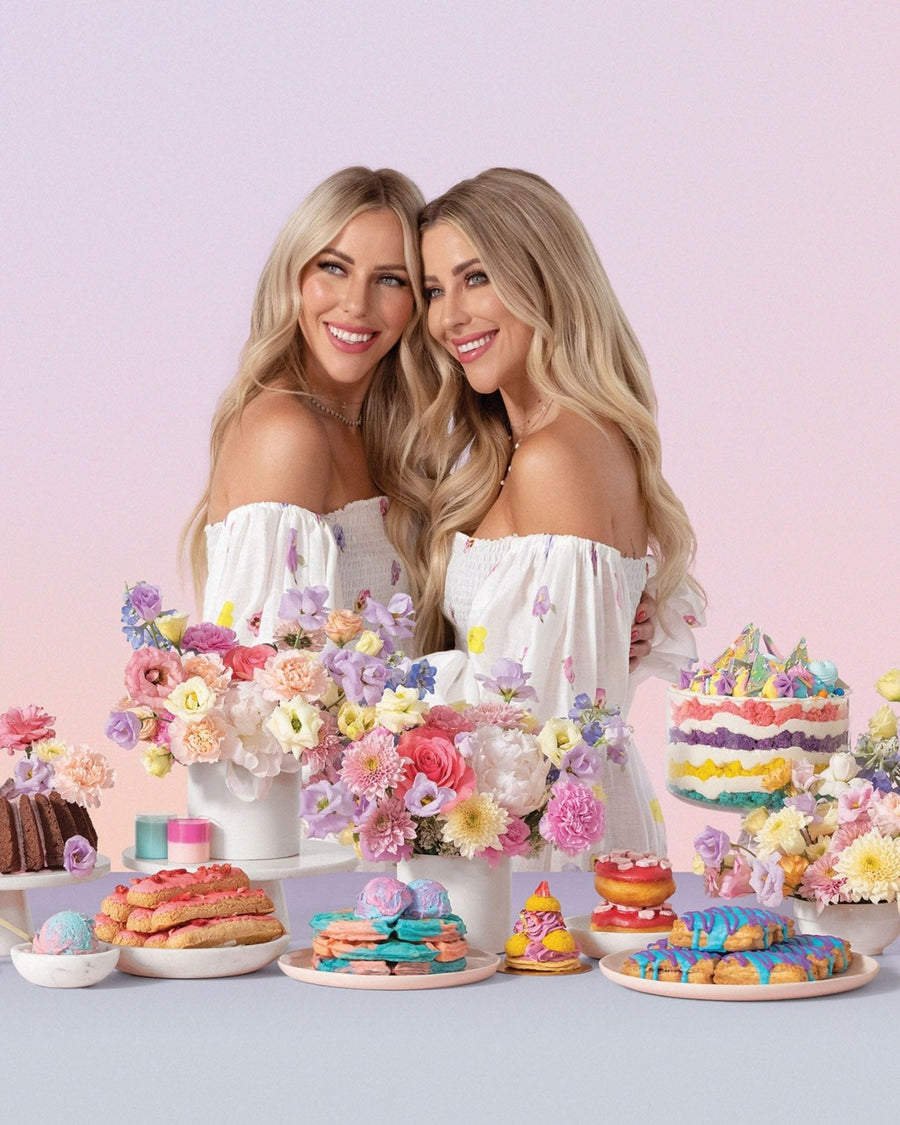 Two smiling women with long blonde hair in white off-shoulder dresses stand together among colorful flowers and vibrant pastries, showcasing the Jenna Rae Cakes at Home Cookbook by Jenna Rae Cakes against a soft pastel backdrop.