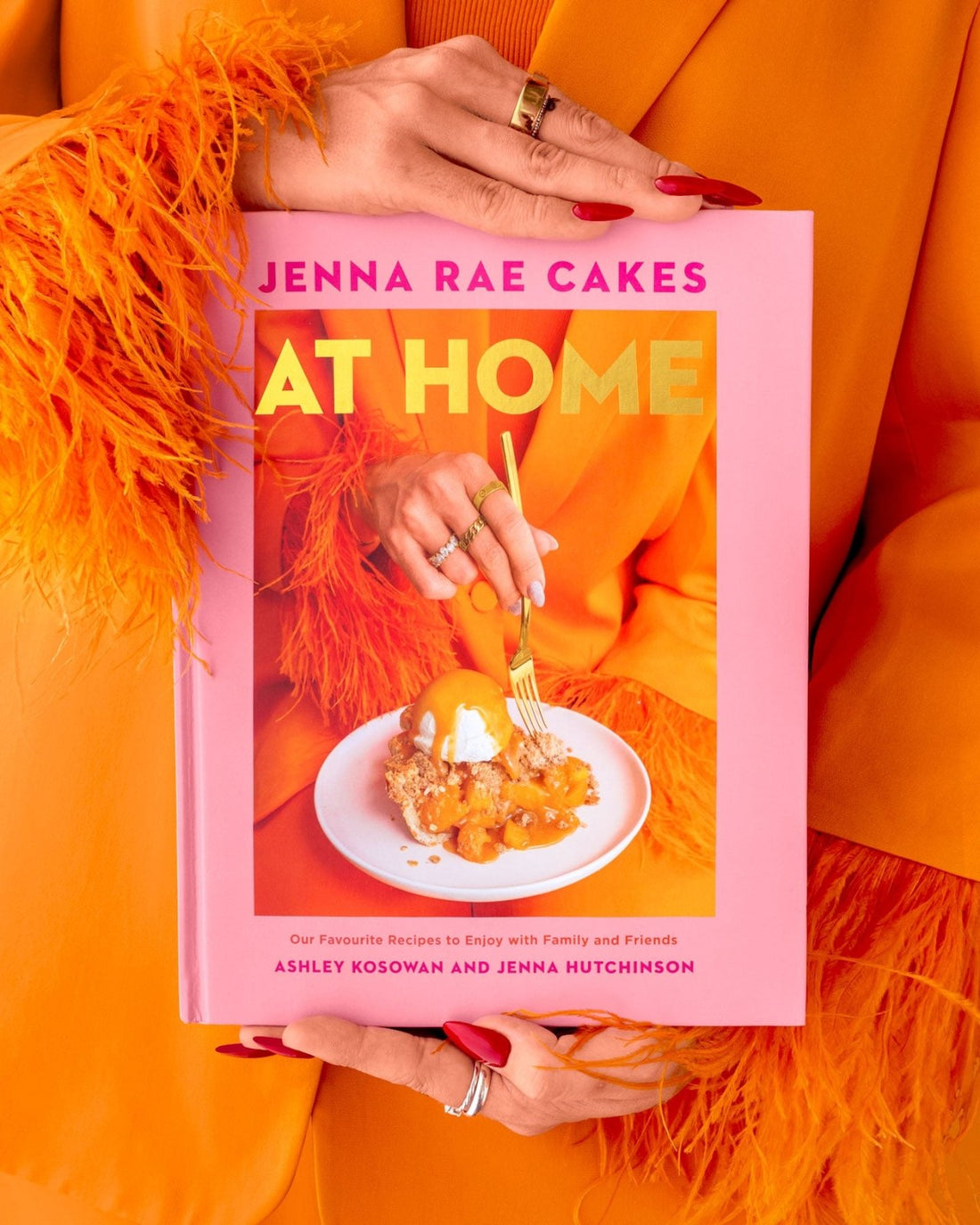 A person in an orange outfit with feathered sleeves holds the Jenna Rae Cakes at Home Cookbook by Jenna Rae Cakes, which features a dessert and gold text on the pink cover. Their nails are painted red and they wear multiple rings.