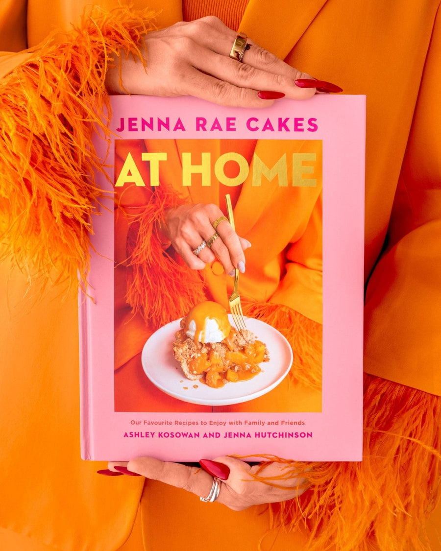 A person in an orange outfit with feathered sleeves holds the Jenna Rae Cakes at Home Cookbook by Jenna Rae Cakes, which features a dessert and gold text on the pink cover. Their nails are painted red and they wear multiple rings.