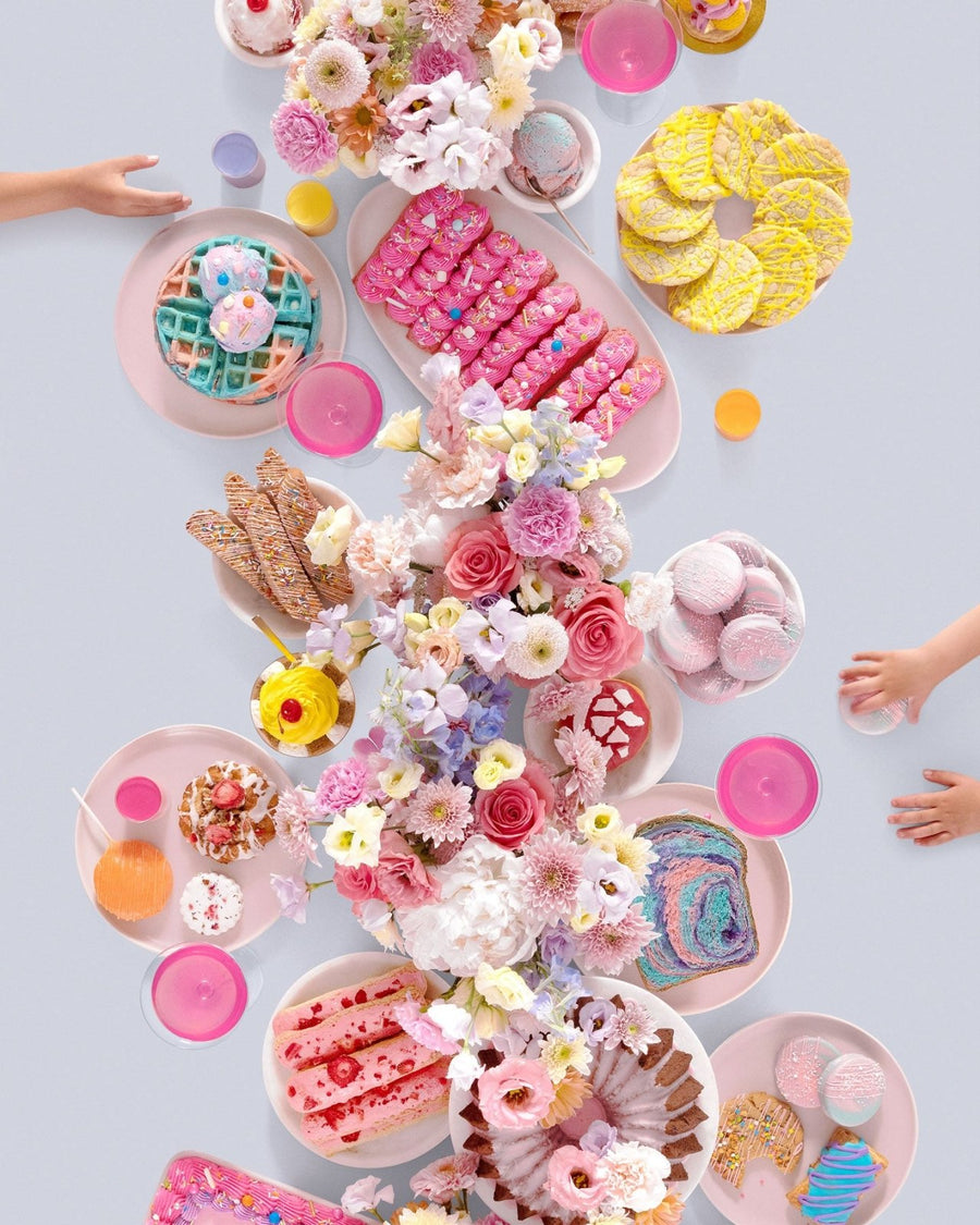 A festive dessert table inspired by Jenna Rae Cakes at Home Cookbook displays pink plates, colorful pastries, waffles, and macarons as children reach for treats. The bright scene captures the playful spirit of Jenna Rae Cakes.