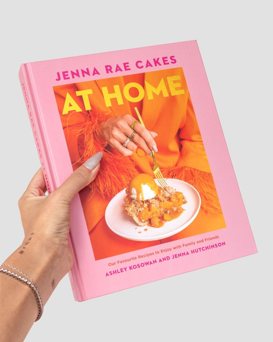 A hand with light nail polish holds the Jenna Rae Cakes at Home Cookbook by Jenna Rae Cakes, featuring a dessert on a plate and a person in an orange outfit using a gold fork on the cover.