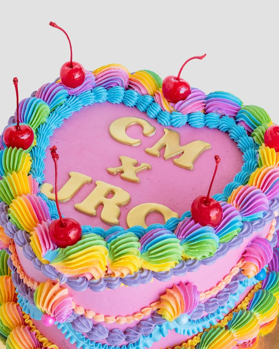 A pink heart-shaped cake from Jenna Rae Cakes, topped with rainbow frosting, red cherries, and yellow letters spelling CM x JRC using the Jenna Rae Cakes x Colour Mill Pack.