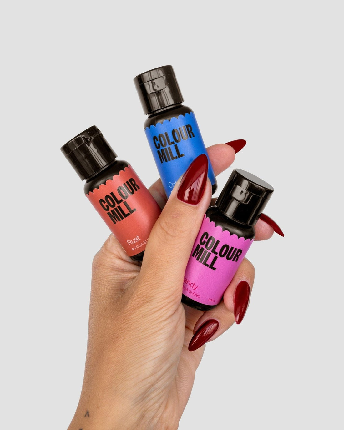 A hand with long red nails holds three Jenna Rae Cakes x Colour Mill Pack bottles from Jenna Rae Cakes in rust, blue, and pink shades against a plain light gray background.