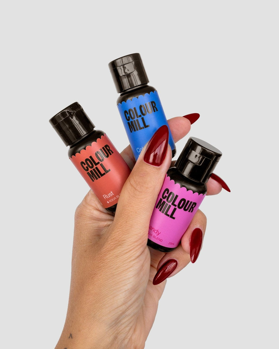 A hand with long red nails holds three Jenna Rae Cakes x Colour Mill Pack bottles from Jenna Rae Cakes in rust, blue, and pink shades against a plain light gray background.