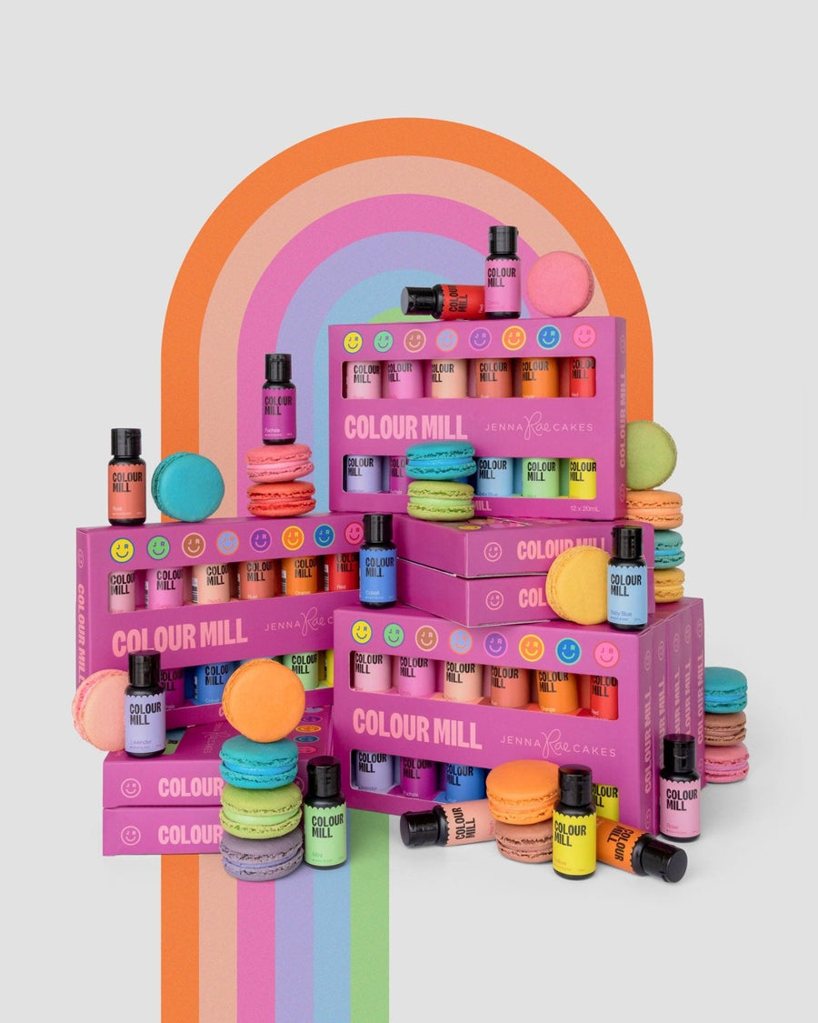 The Jenna Rae Cakes x Colour Mill Pack by Jenna Rae Cakes is displayed alongside colorful macarons before a rainbow arch, featuring vibrant, playful energy and bright pink packaging on a light background.